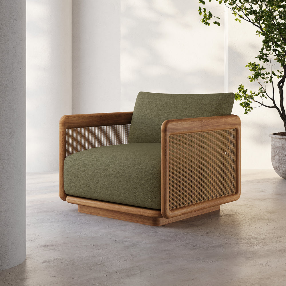 Lemieux et Cie Dock Teak Lounge Chair with olive cushions and woven cane panels, outdoor lounge chair on concrete floor in dappled natural light.