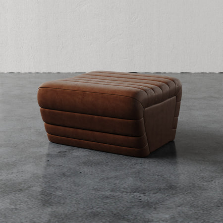 Lemieux et Cie Brunoy Ottoman, channel-tufted leather ottoman in warm cognac tones, shown on polished concrete floor against textured plaster walls.