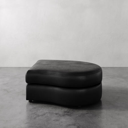 Lemieux et Cie Rulmann Right Ottoman in black leather, low-profile cushioned ottoman on concrete floor against neutral plaster walls.