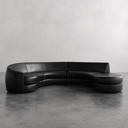 Lemieux et Cie Rulmann sectional in black leather, curved four-piece sofa with rounded ottoman, shown on concrete floor against neutral walls.