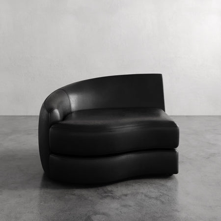 Lemieux et Cie Rulmann Left Arm Chair in black leather, sculptural low-profile seating with rounded back, shown on concrete floor against white wall.