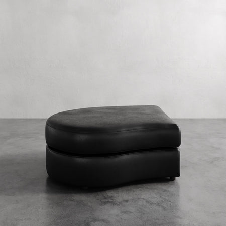 Lemieux et Cie Rulmann Left Ottoman in black leather, low-profile curved ottoman on concrete floor against neutral plaster wall.