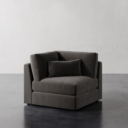 Lemieux et Cie Lacroix Corner Chair in charcoal upholstery, modular sectional piece with bolster pillow, shown on dark concrete floor against textured plaster walls.