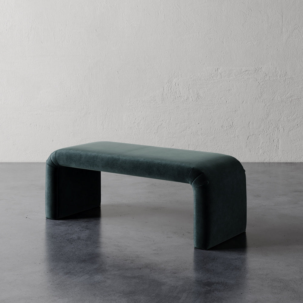 Lemieux et Cie Nadia 48" Bench in deep teal upholstery, sculptural bench with curved waterfall form, shown on concrete floor against textured plaster wall.