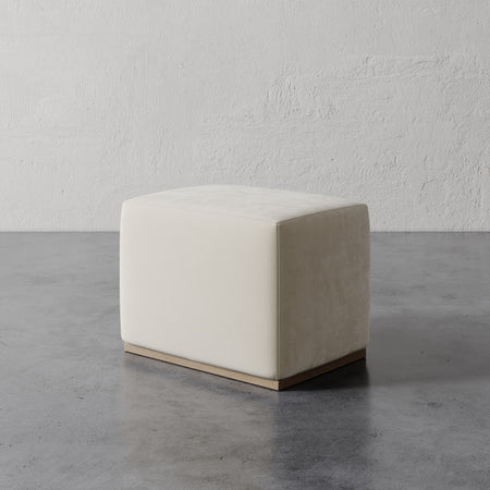 Lemieux et Cie Sophie Rectangular Ottoman in ivory upholstery with taupe side panel, low-profile ottoman on stone flooring against textured plaster walls.