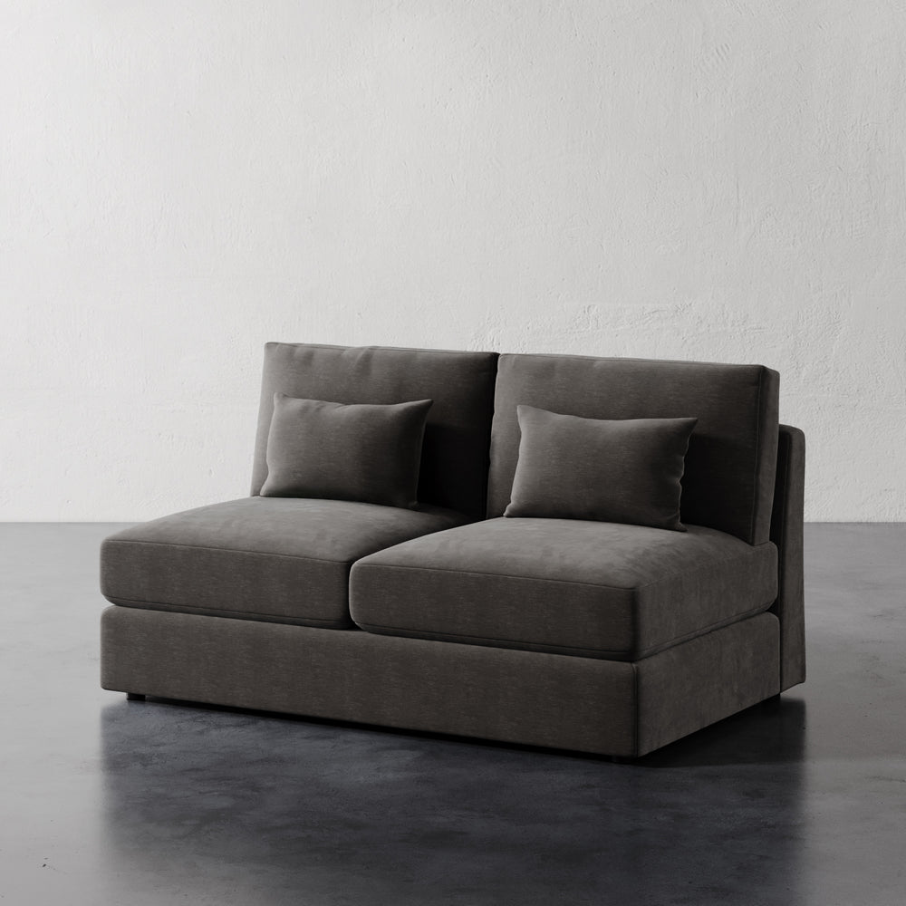 Lemieux et Cie Lacroix Armless Loveseat in charcoal gray, modular two-seat sofa with low profile and bolster pillows, on dark stone floor against textured plaster walls.