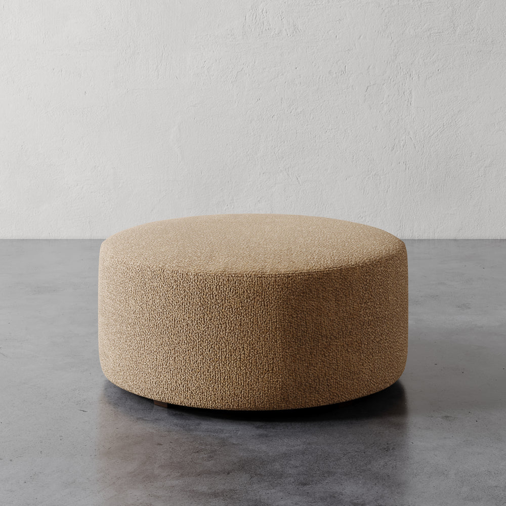 Lemieux et Cie Fabien 36" Round Ottoman, low-profile cylindrical ottoman in warm textured upholstery, shown on gray concrete floor against white plaster wall.