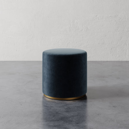 Lemieux et Cie Sophie Round Ottoman in dark upholstery with brass base, cylindrical ottoman on concrete floor against textured plaster wall.