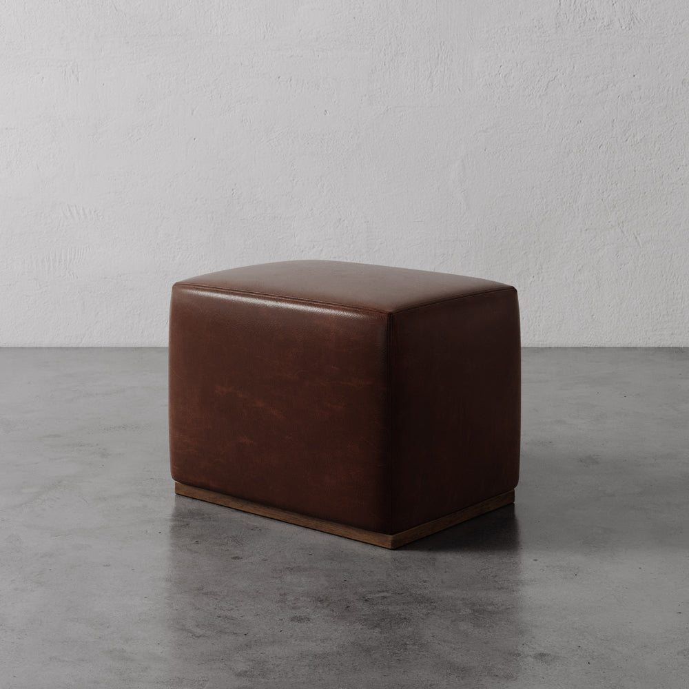 Lemieux et Cie Sophie Leather Rectangular Ottoman in warm brown leather, low-profile ottoman with wood plinth base, shown on concrete floor against textured plaster walls.