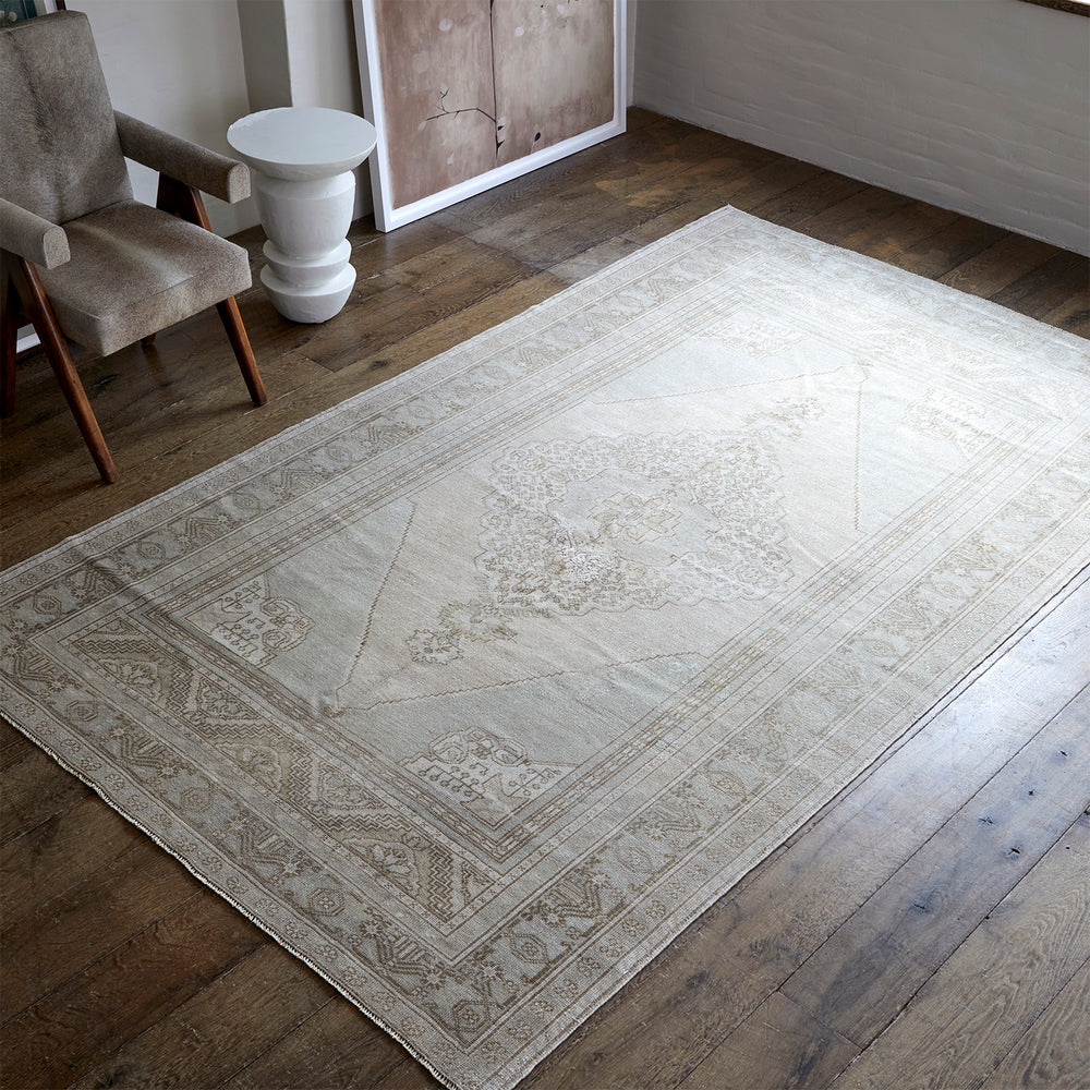 Lemieux et Cie Anatolian Village Rug in ivory and taupe, vintage geometric medallion pattern, shown on wood floors beside upholstered chair and plaster pedestal table.