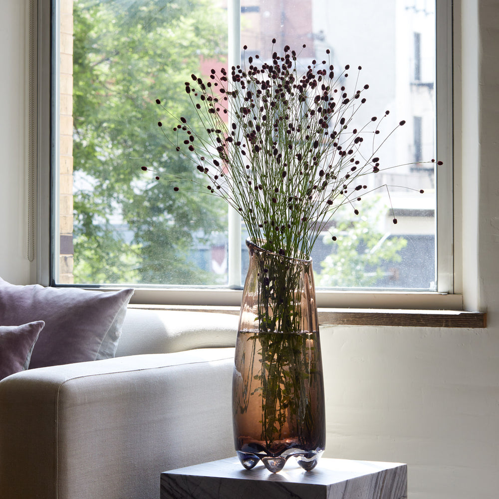 Lemieux et Cie Fleur de Verre vase with organic flowing form, transparent glass, styled on neutral sofa with dried botanicals by sunlit window.