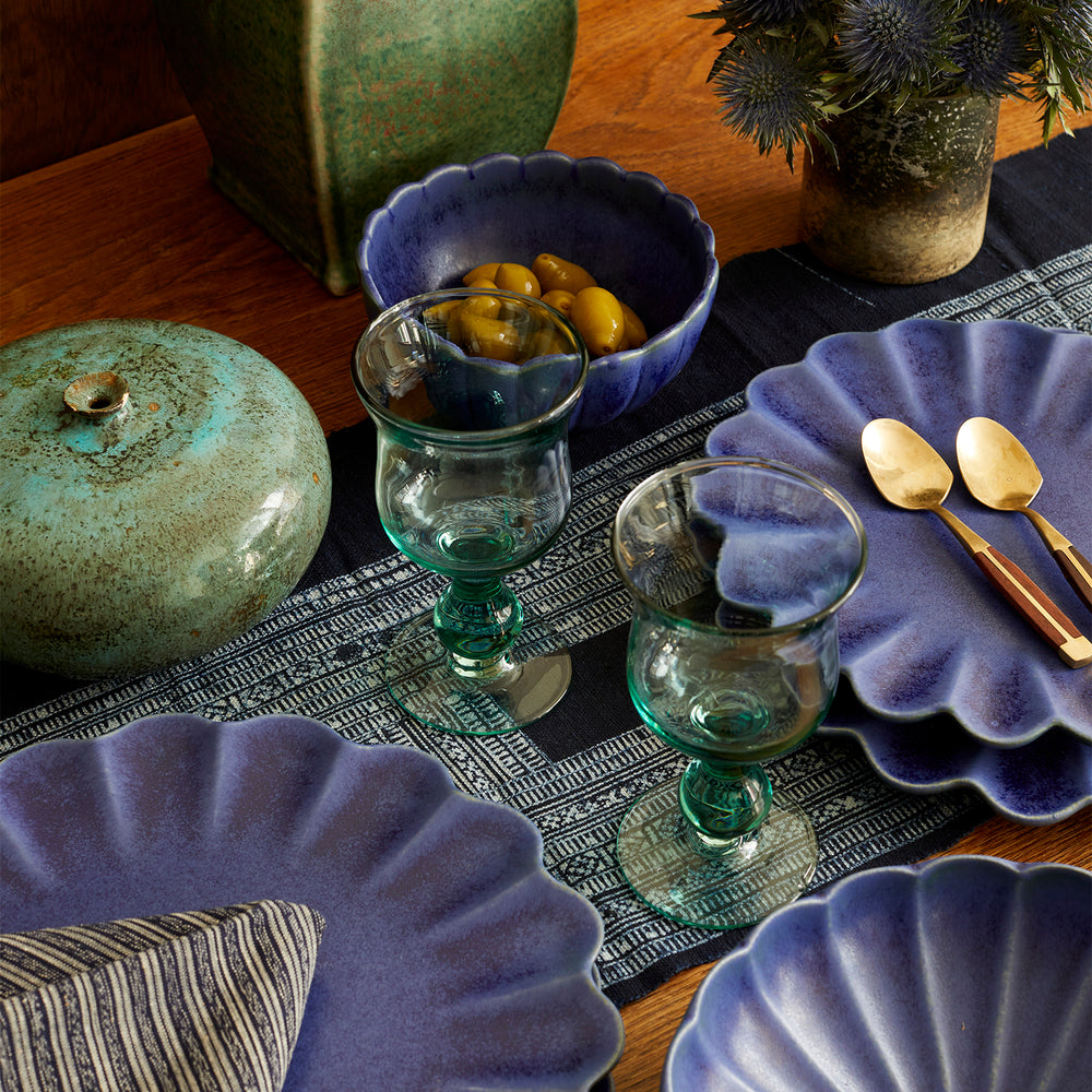 Lemieux et Cie Dhalia Stoneware dinnerware set in blue with scalloped edges, styled on wood table with green ceramic jar, glassware and woven runner.
