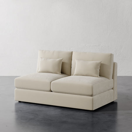 Lemieux et Cie Lacroix Armless Loveseat in ivory, modular sofa with tailored cushions and bolster pillows on dark concrete floor against textured plaster walls.