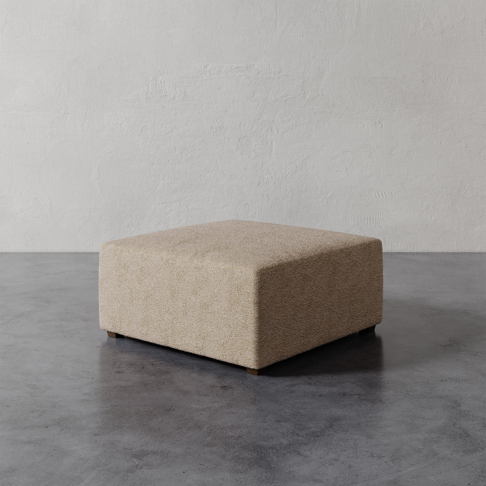 Lemieux et Cie Fabien 36" Square Ottoman in neutral upholstery, low-profile ottoman on dark concrete floor against textured plaster walls.