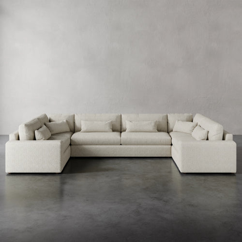 Lemieux et Cie Lacroix U-Sectional in neutral upholstery, modular sofa with rectilinear form and bolster pillows, shown on dark concrete floors against textured plaster walls.