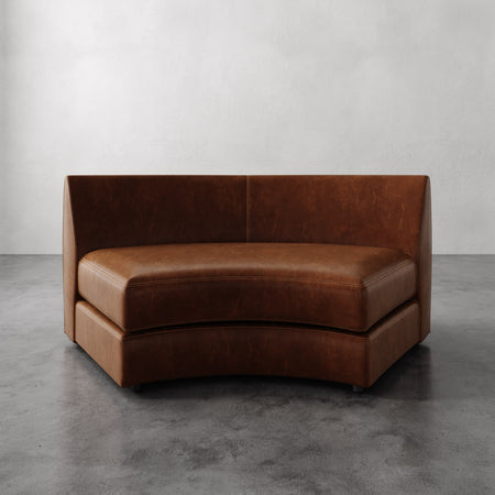 Lemieux et Cie Rulmann Wedge in warm cognac leather, curved banquette seating with high back, shown on concrete floor against white walls.