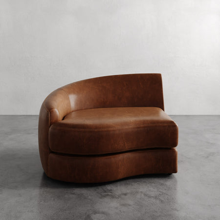 Lemieux et Cie Rulmann Left Arm Chair in warm cognac leather, curved sectional piece on concrete floor against neutral plaster walls.