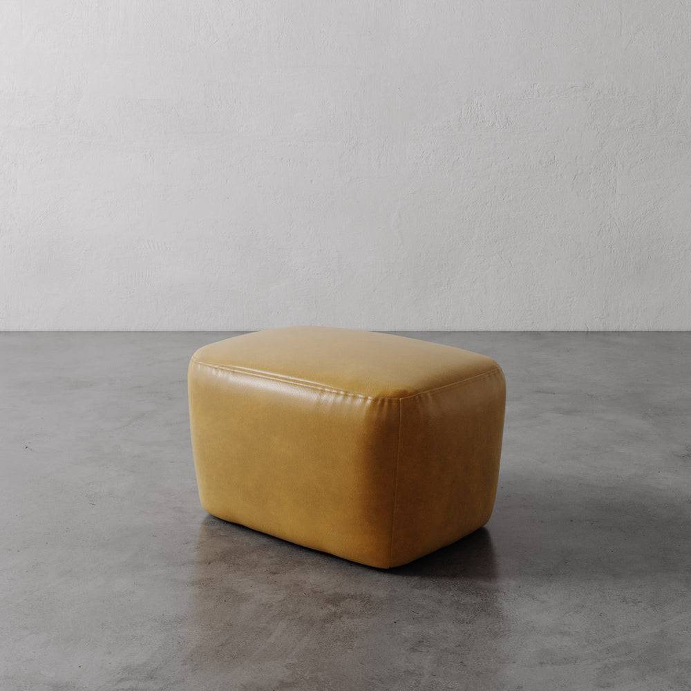 Lemieux et Cie Aurélie Leather Ottoman in warm tobacco leather, softly rounded ottoman with sculptural form, shown on concrete floor against neutral plaster walls.
