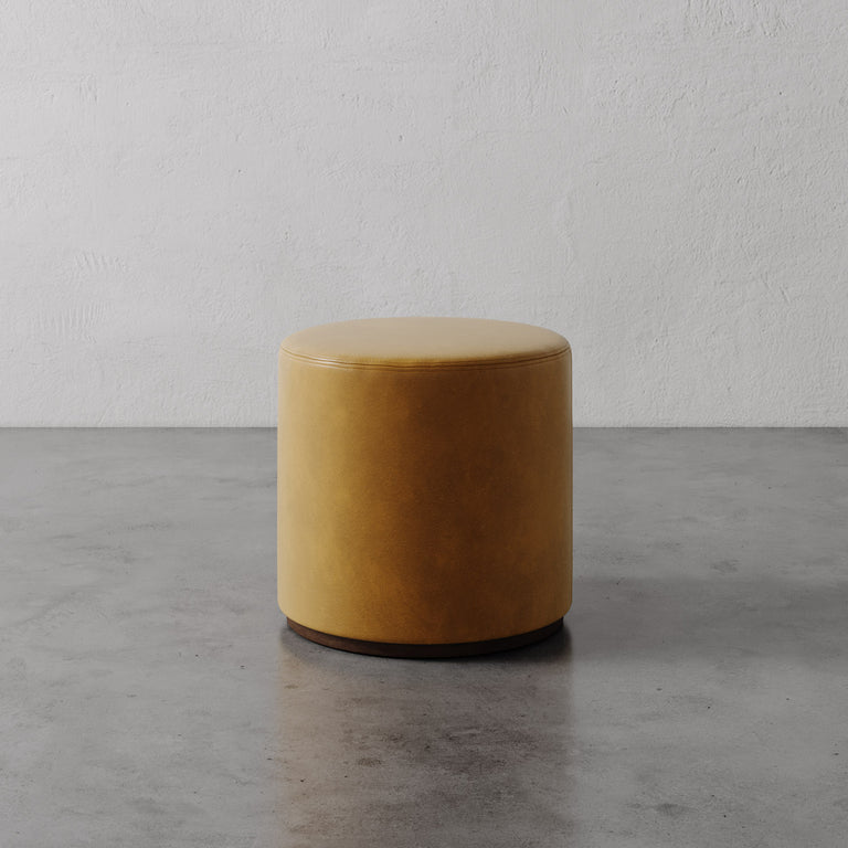 Lemieux et Cie Sophie Round Ottoman in warm gold leather, cylindrical ottoman with dark base, on concrete floor against textured plaster wall.