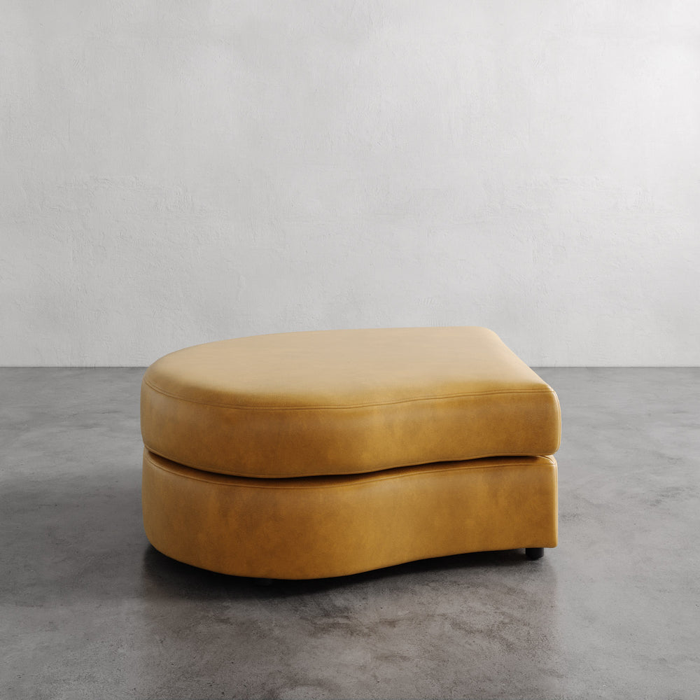 Lemieux et Cie Rulmann Left Ottoman in warm tobacco, rounded low-profile form, shown on polished concrete floor against neutral plaster walls.