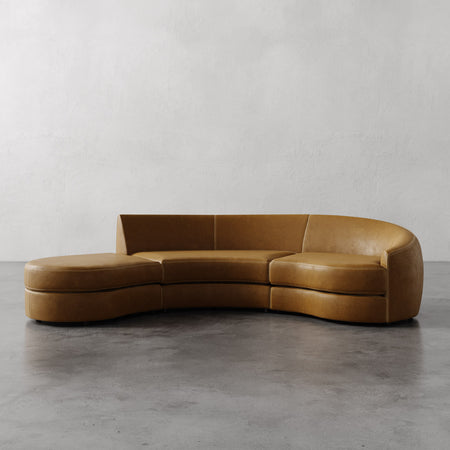 Lemieux et Cie Rulmann Sectional in warm tan leather, curved low-profile sofa on polished concrete floor against textured white walls.