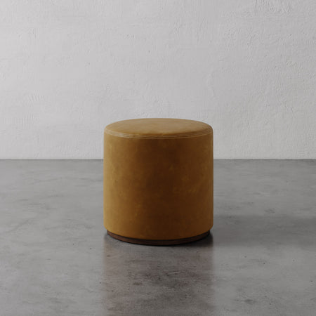 Lemieux et Cie Sophie Round Ottoman in warm tobacco tone, cylindrical ottoman on dark base, shown on concrete floor against textured plaster wall.