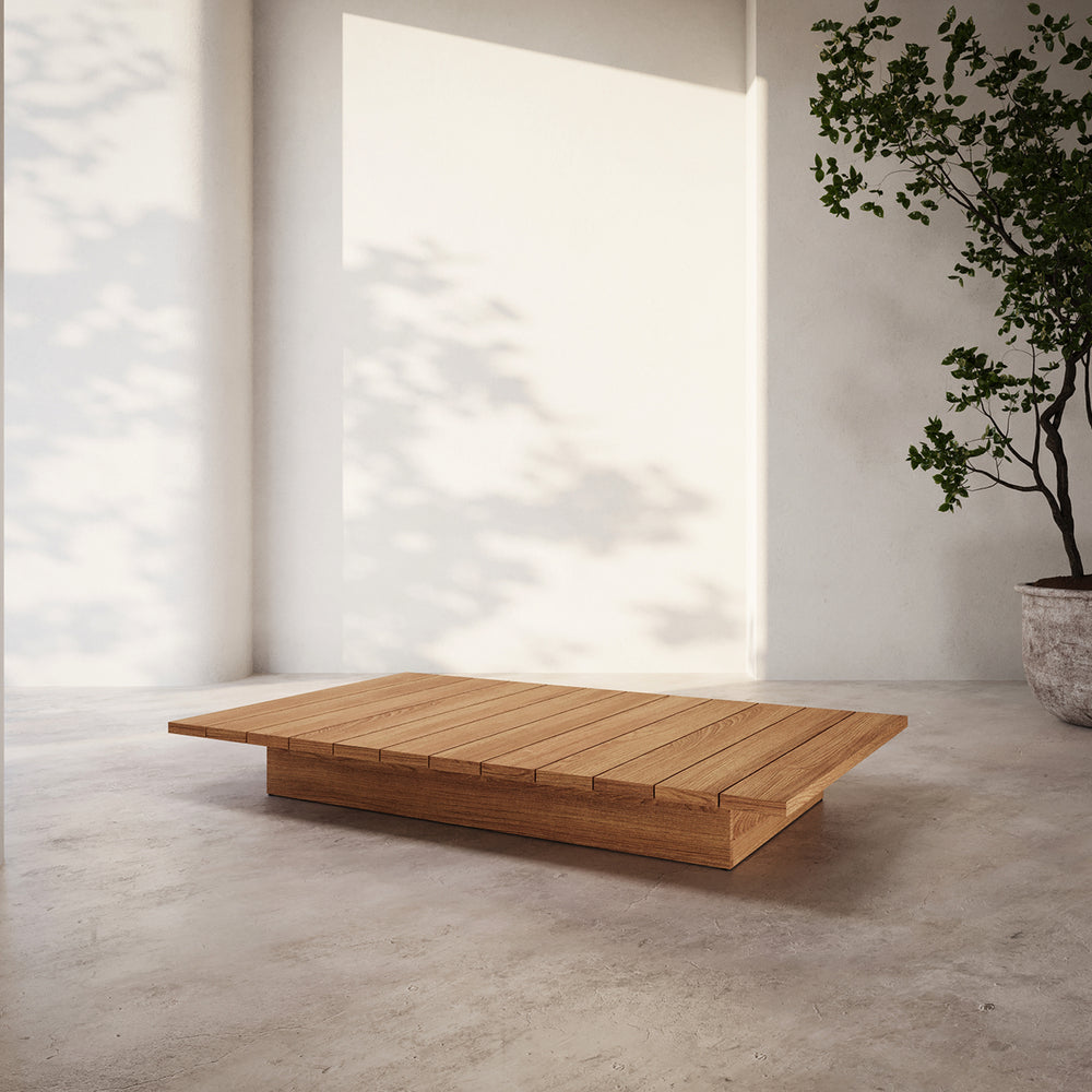 Lemieux et Cie S2 Teak Rectangular Coffee Table in solid teak with slatted top, low-profile outdoor table on concrete floor in white-walled room with dappled sunlight.