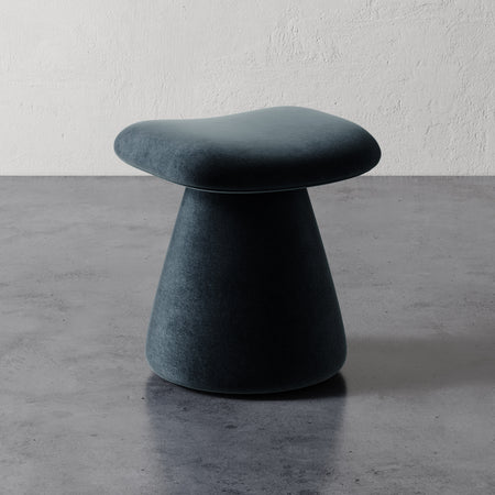 Lemieux et Cie Romy Ottoman in dark teal upholstery, sculptural pedestal stool with flared base and organic seat, shown on polished concrete floor against textured plaster wall.