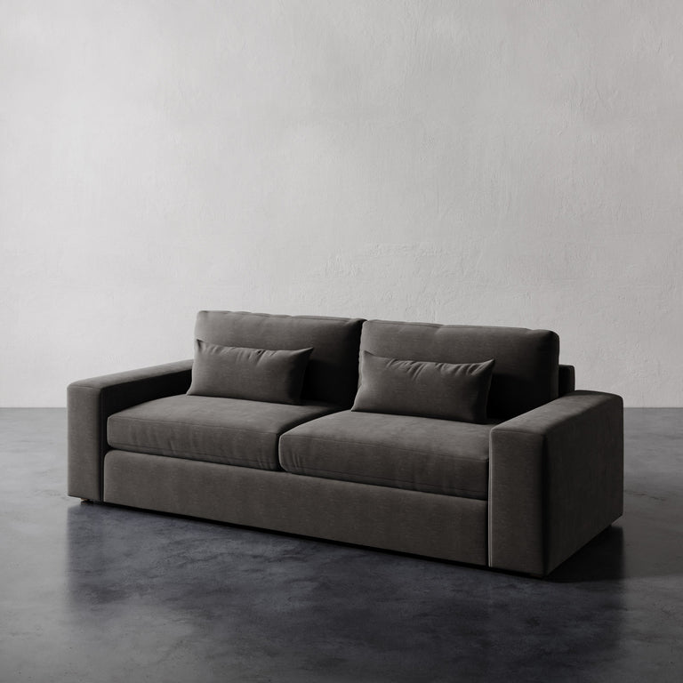 Lemieux et Cie Lacroix Sofa in charcoal upholstery, geometric two-seat sofa with bolster pillows, shown on dark concrete floor against textured plaster walls.
