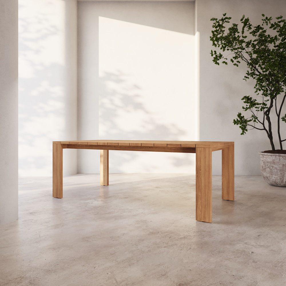 Lemieux et Cie S2 Teak Rectangular Dining Table in solid teak wood, outdoor dining table with chunky legs shown on concrete floor in sunlit white-walled room.