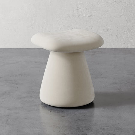 Lemieux et Cie Romy Ottoman in ivory, sculptural stool with flared pedestal base and organic contoured seat, on concrete floor against textured plaster wall.