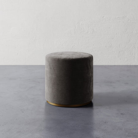 Lemieux et Cie Sophie Round Ottoman in charcoal upholstery with brass base, cylindrical ottoman shown on concrete floor against textured plaster wall.