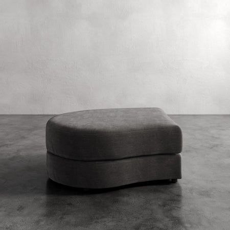 Lemieux et Cie Rulmann Left Ottoman in charcoal, rounded low-profile ottoman on polished concrete floor against textured plaster wall.