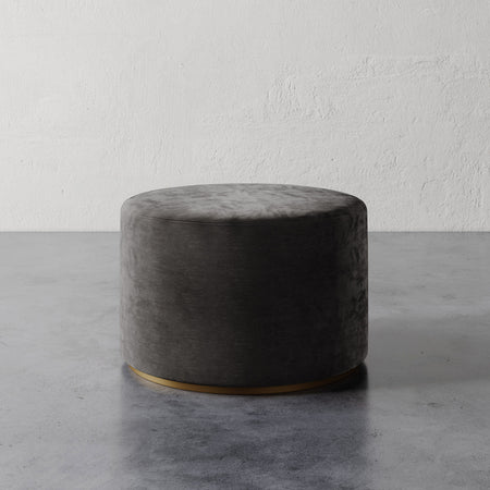Lemieux et Cie Sophie Round Ottoman in charcoal upholstery with brass base detail, cylindrical ottoman shown on concrete floor against textured plaster wall.
