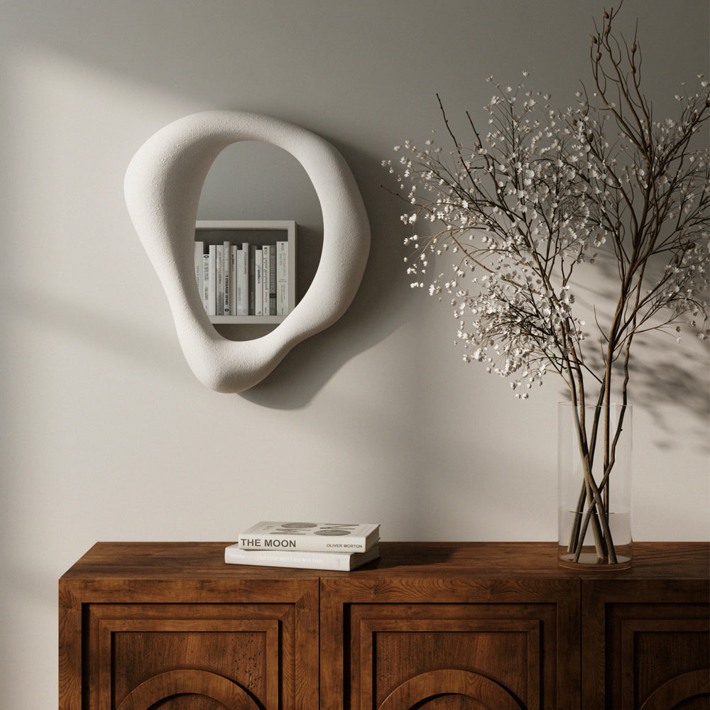 Lemieux et Cie hand-sculpted mirror with organic asymmetrical frame, wall-mounted shelf displaying books, styled atop warm wood cabinet with neutral plaster walls and delicate branches in glass vase.