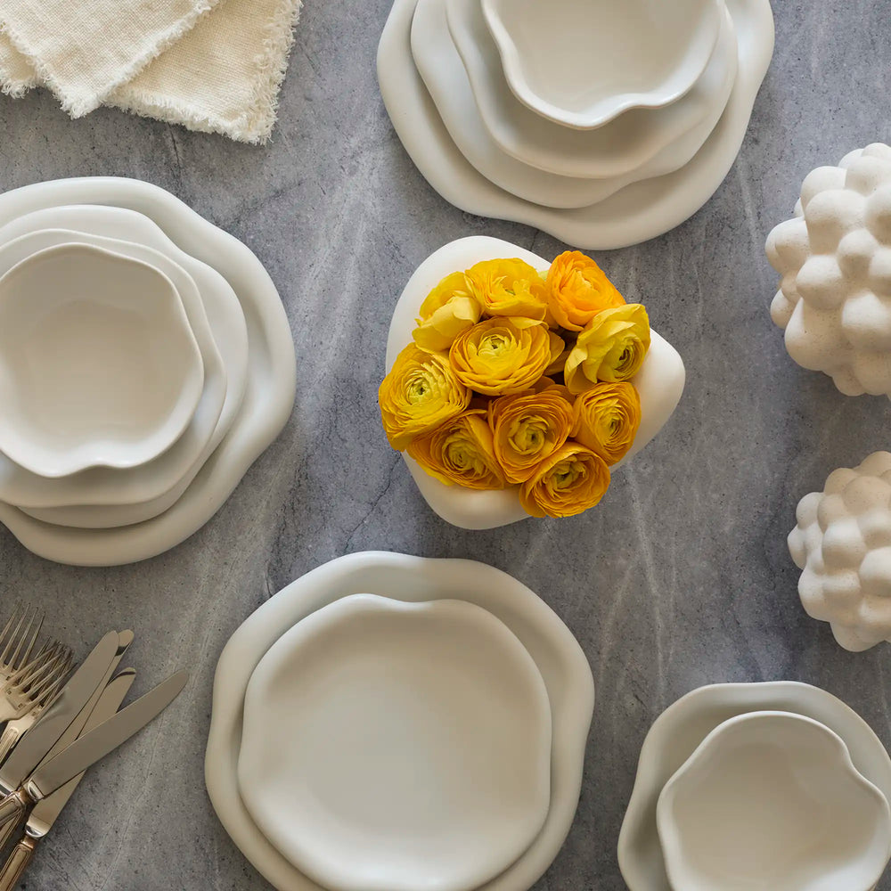 Lemieux et Cie Amorphous Dinner Plate Set in soft white matte stoneware, organic-edged dinnerware shown on grey stone surface with yellow ranunculus and sculptural vases.