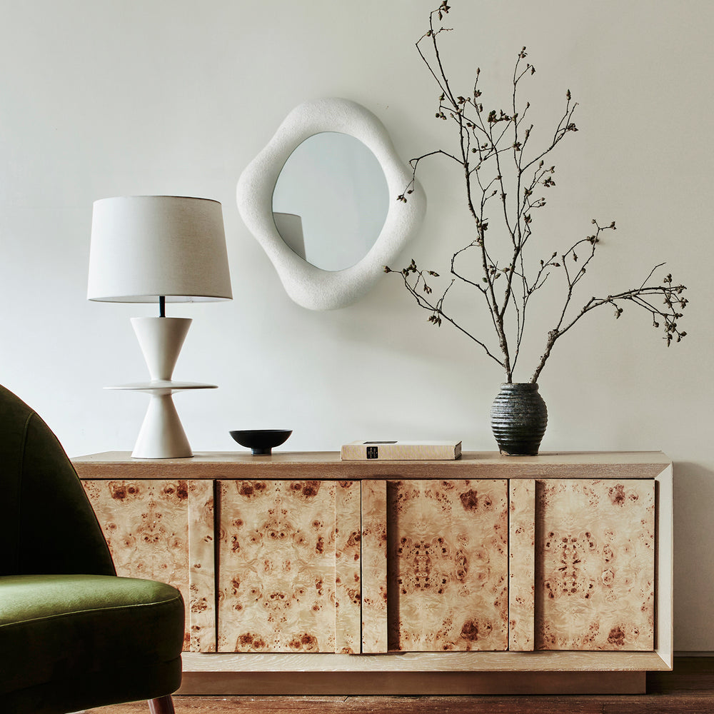Lemieux et Cie hand-sculpted mirror with asymmetrical organic frame, styled above burl wood credenza with table lamp and dried branches on neutral wall.