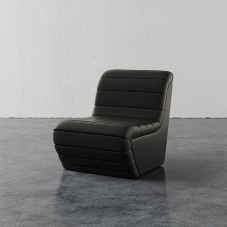 Lemieux et Cie Brunoy Leather Chair in dark leather with horizontal channel tufting, sculptural slipper chair on concrete floor against white plaster wall.
