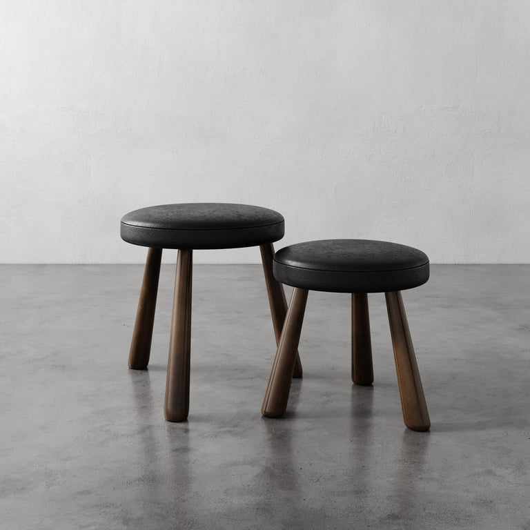 Lemieux et Cie Madeleine Leather Stool in dark grey leather with tapered legs, two stools on polished concrete floor against white plaster wall.