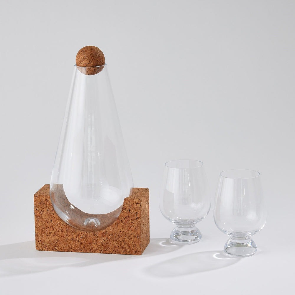Lemieux et Cie glass decanter with cork stopper and geometric cork base, accompanied by two stemmed glasses on white studio backdrop.