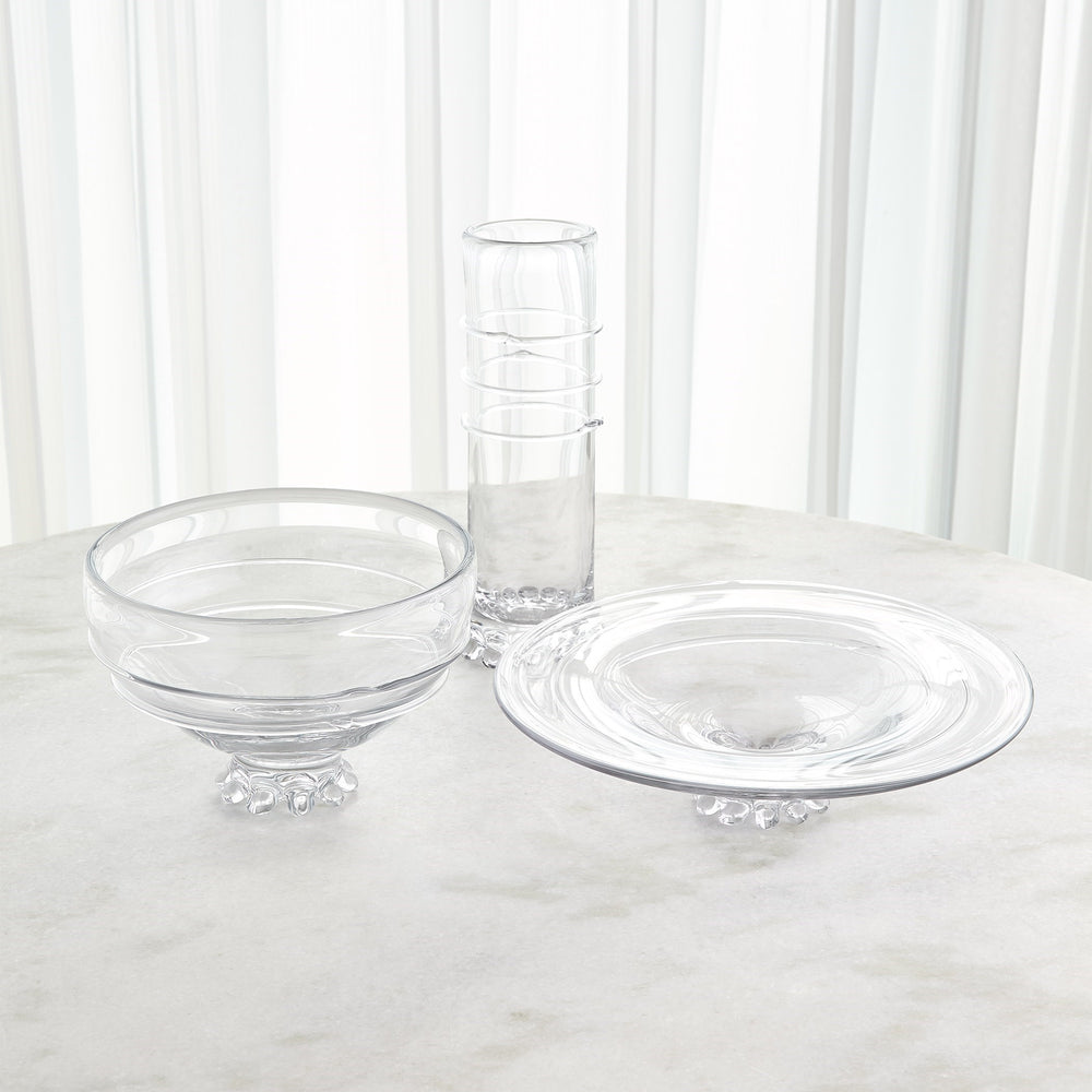 Lemieux et Cie Verre Lisse Collection glass bowl, plate and vase with sculptural footed bases, arranged on marble surface before white drapery.