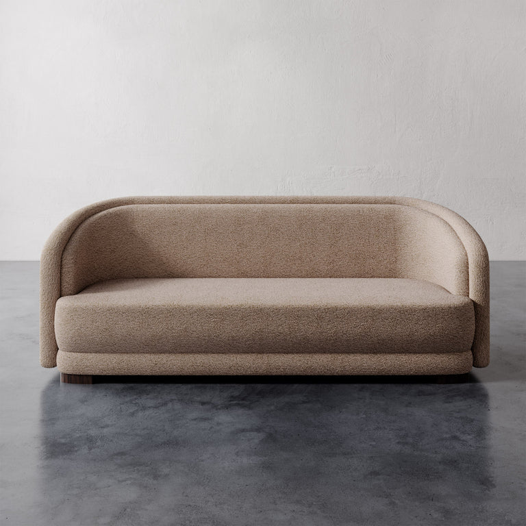Lemieux et Cie Valence Sofa in warm taupe, low-profile rounded form, shown on dark concrete flooring against neutral plaster walls.