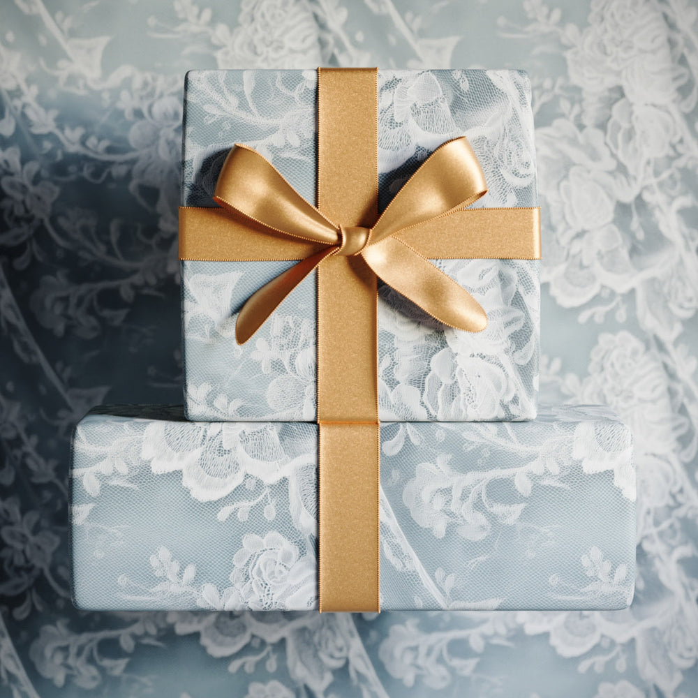 Lemieux et Cie Chantilly Lace Gift Wrapping Paper in ivory and soft blue, stacked boxes tied with gold ribbon on a matching surface.