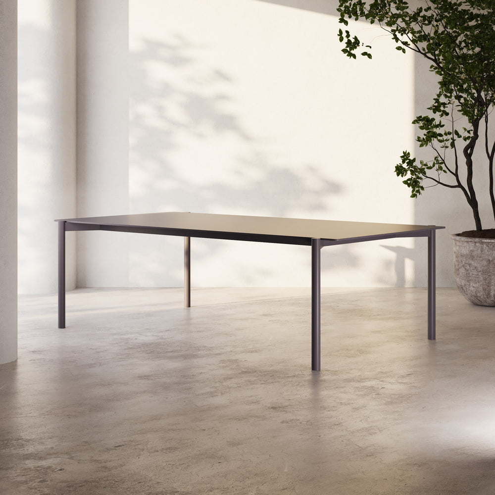 Lemieux et Cie Hudson 84" Rectangular Dining Table with slim dark frame and light top, dining table on concrete floor in sun-dappled room with potted tree.
