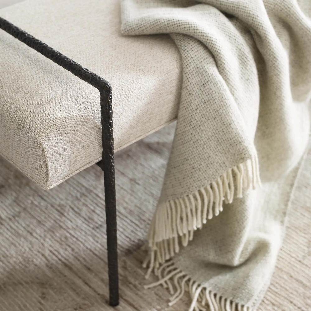 Lemieux et Cie Imane Bench in neutral upholstery with deeply hammered bronze frame, close view of textured throw draped over bench, light wood flooring.