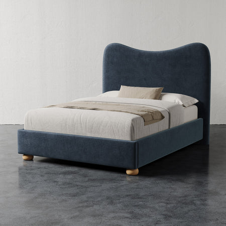 Lemieux et Cie Danielle Bed in dark blue upholstery with sculptural curved headboard and rounded feet, styled with neutral bedding on stone floor.