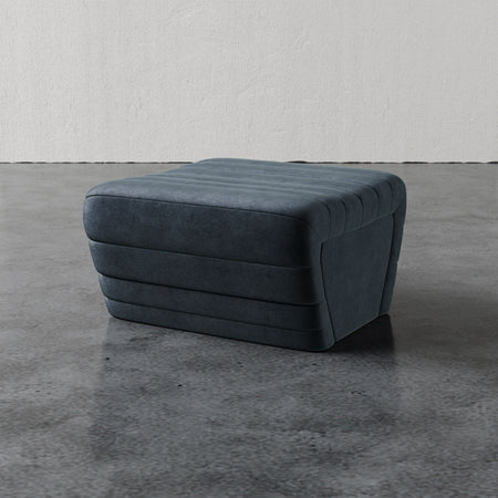 Lemieux et Cie Brunoy Ottoman in charcoal with channel-tufted detail, upholstered ottoman on polished concrete floor against textured plaster walls.