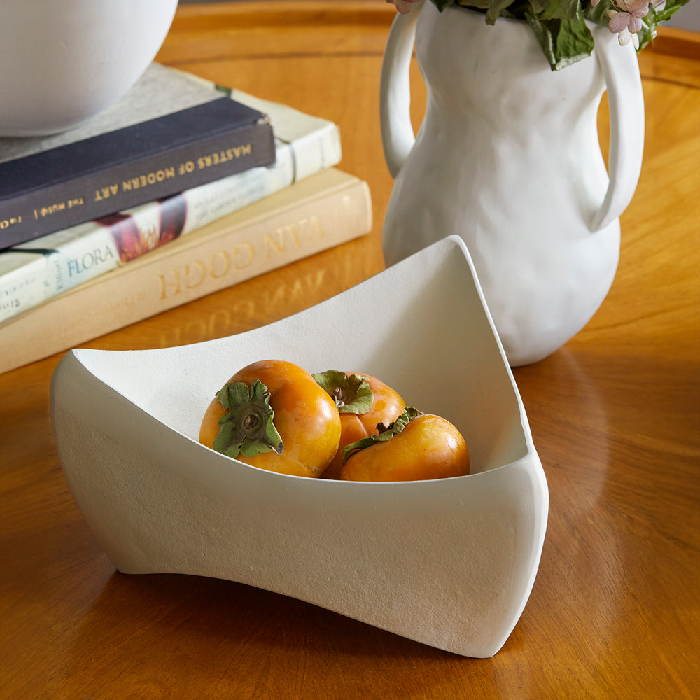Lemieux et Cie Marius Square Bowl in white, sculptural ceramic bowl with angular origami-inspired form, styled with persimmons on wood table beside stacked books and vase.