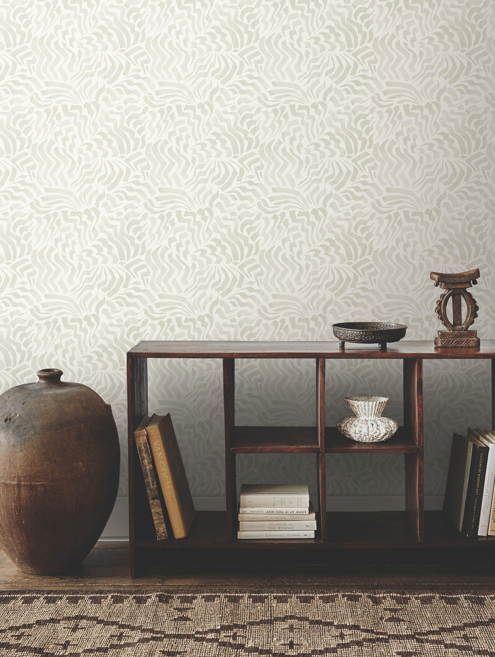 Lemieux et Cie Zora Waves wallpaper in light gray on creamy white, wave-pattern wall covering shown in a sitting room with dark wood shelving and woven rug.