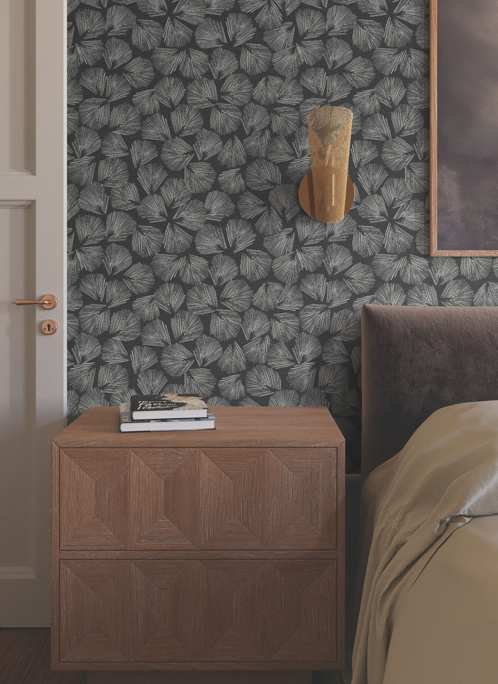Lemieux et Cie Elora Leaf Wallpaper in sage green with metallic veining, botanical pattern wallcovering, styled in a bedroom with textured nightstand and warm sconce lighting.